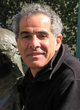 Outdoor headshot of a 55 year old man with olive skin and curly salt and pepper hair smiling